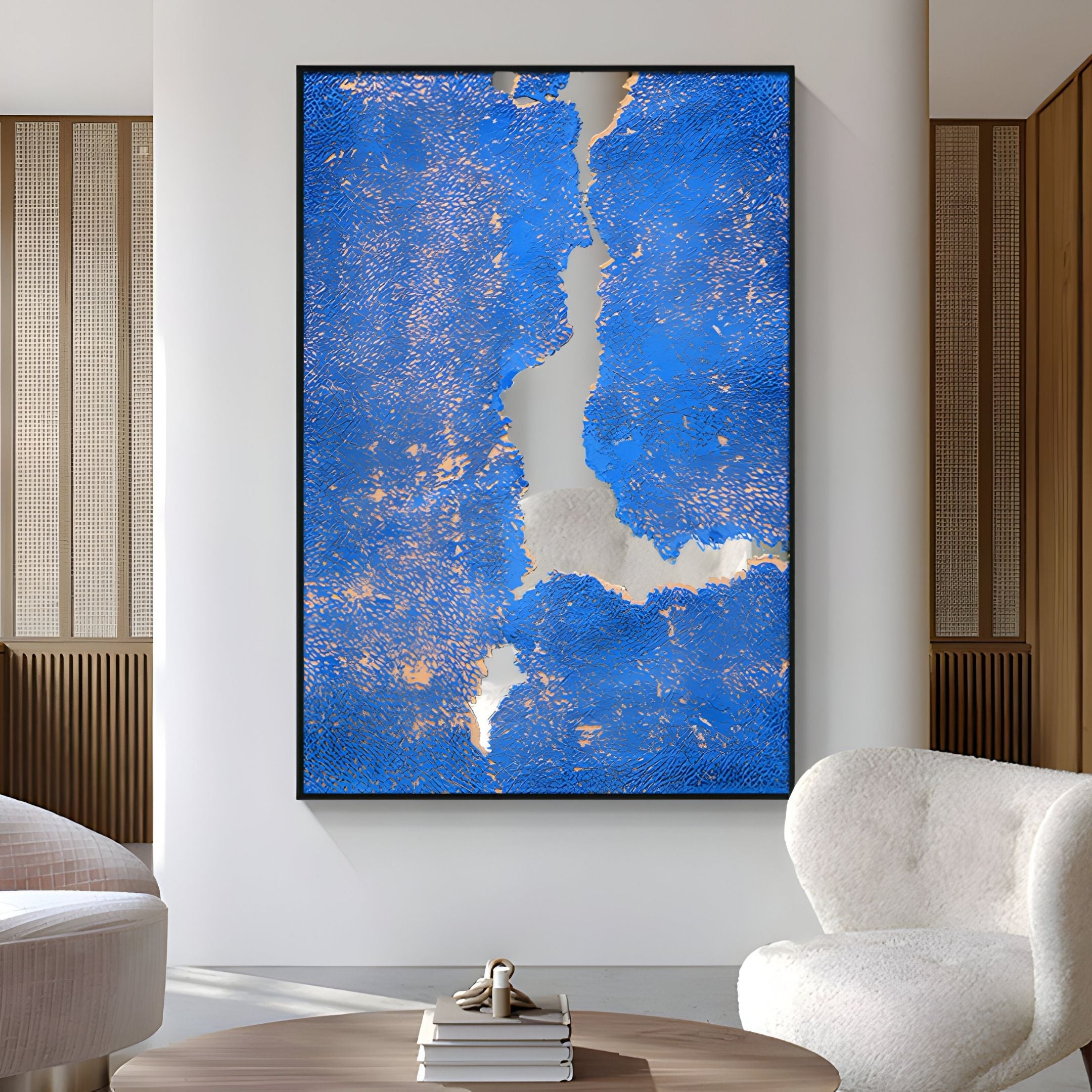 A Cerulex Blue Rift Textured Steel Mirror 3D Wall Art by Giant Sculptures, featuring bold blue textures and gold accents, is displayed on a white wall in a modern living room with white chairs and a small table.