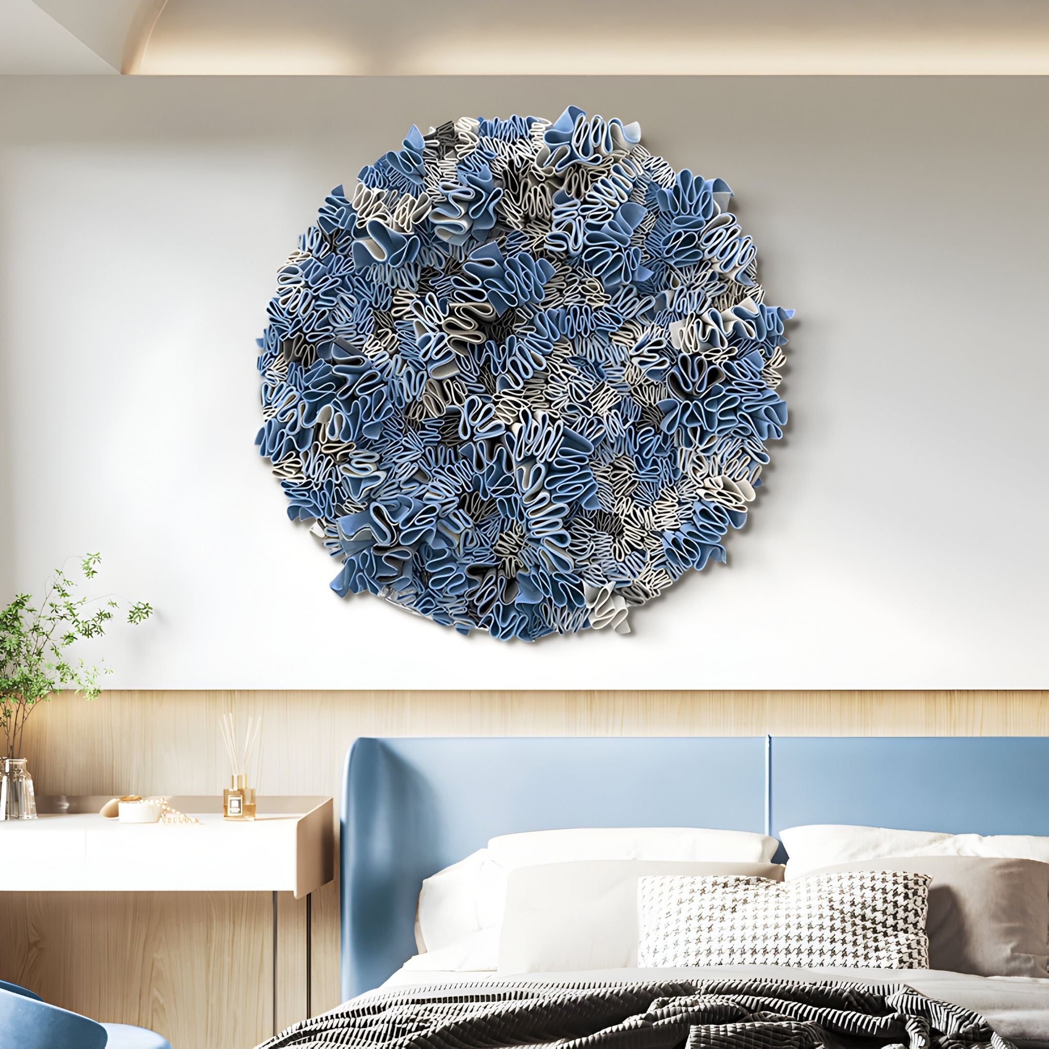 The Rivora Blue Ribbon Leather Circular 3D Wall Art by Giant Sculptures hangs above a bed with matching bedding in a modern, well-lit bedroom. On the left, a small desk with a plant enhances the contemporary décor.