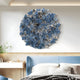 The Rivora Blue Ribbon Leather Circular 3D Wall Art by Giant Sculptures hangs above a bed with matching bedding in a modern, well-lit bedroom. On the left, a small desk with a plant enhances the contemporary décor.