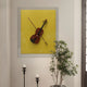 The Archetto Yellow Violin Acrylic Framed 3D Wall Art by Giant Sculptures features a violin and bow on a bold yellow background, making striking modern wall decor above black candle holders and a leafy green potted plant.