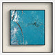 The Trevasse Aqua Blue Textured Abstract Framed Wall Art by Giant Sculptures features a teal blue background with white splashes, black speckles, and silver streaks in a black shadow box frame displayed on a light beige wall.