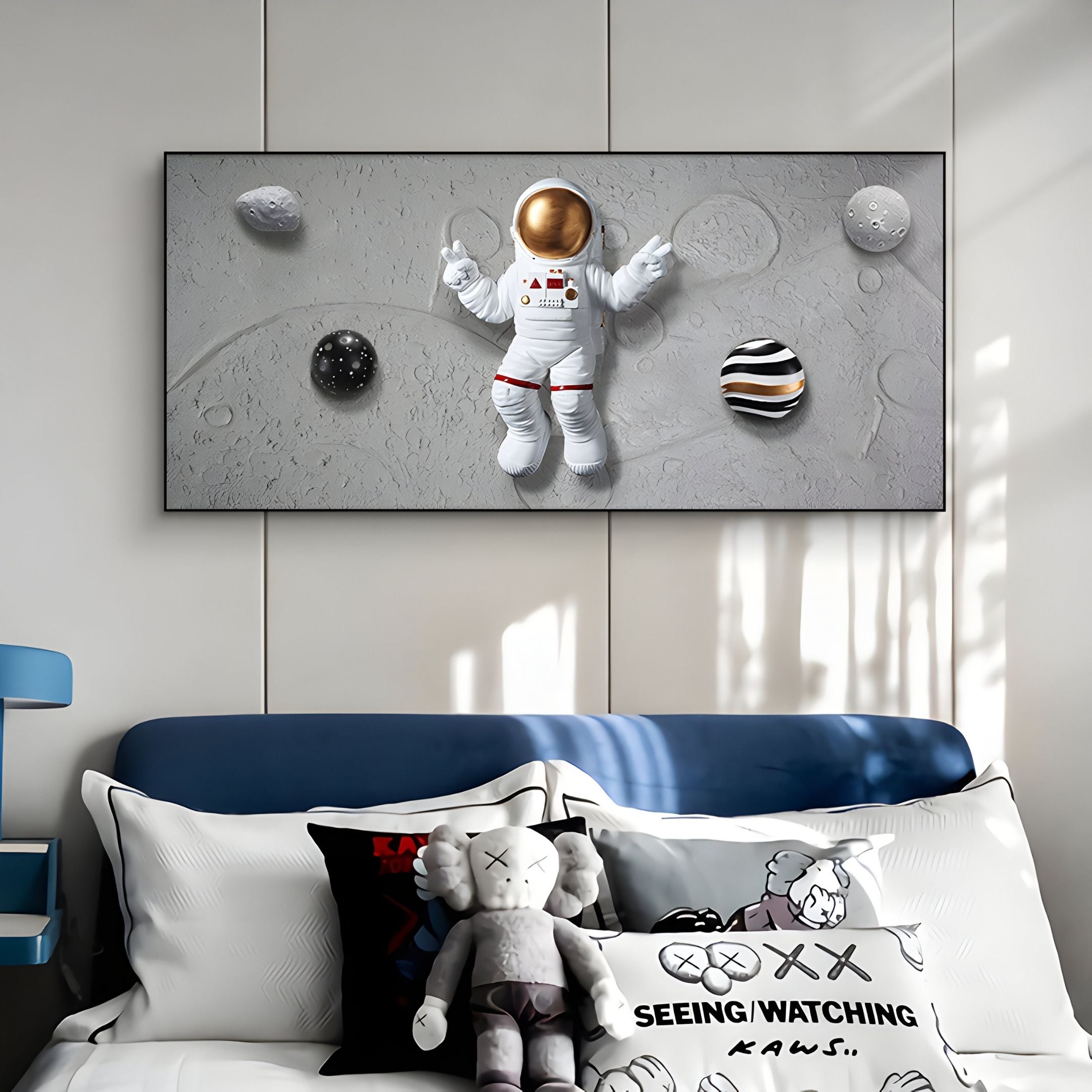 A bedroom with blue and white decor features a bed with plush toys and KAWS pillows. Above, the Astravera Galactic Orbit Planet Astronaut 3D Wall Art by Giant Sculptures adds a perfect touch of space-themed style.