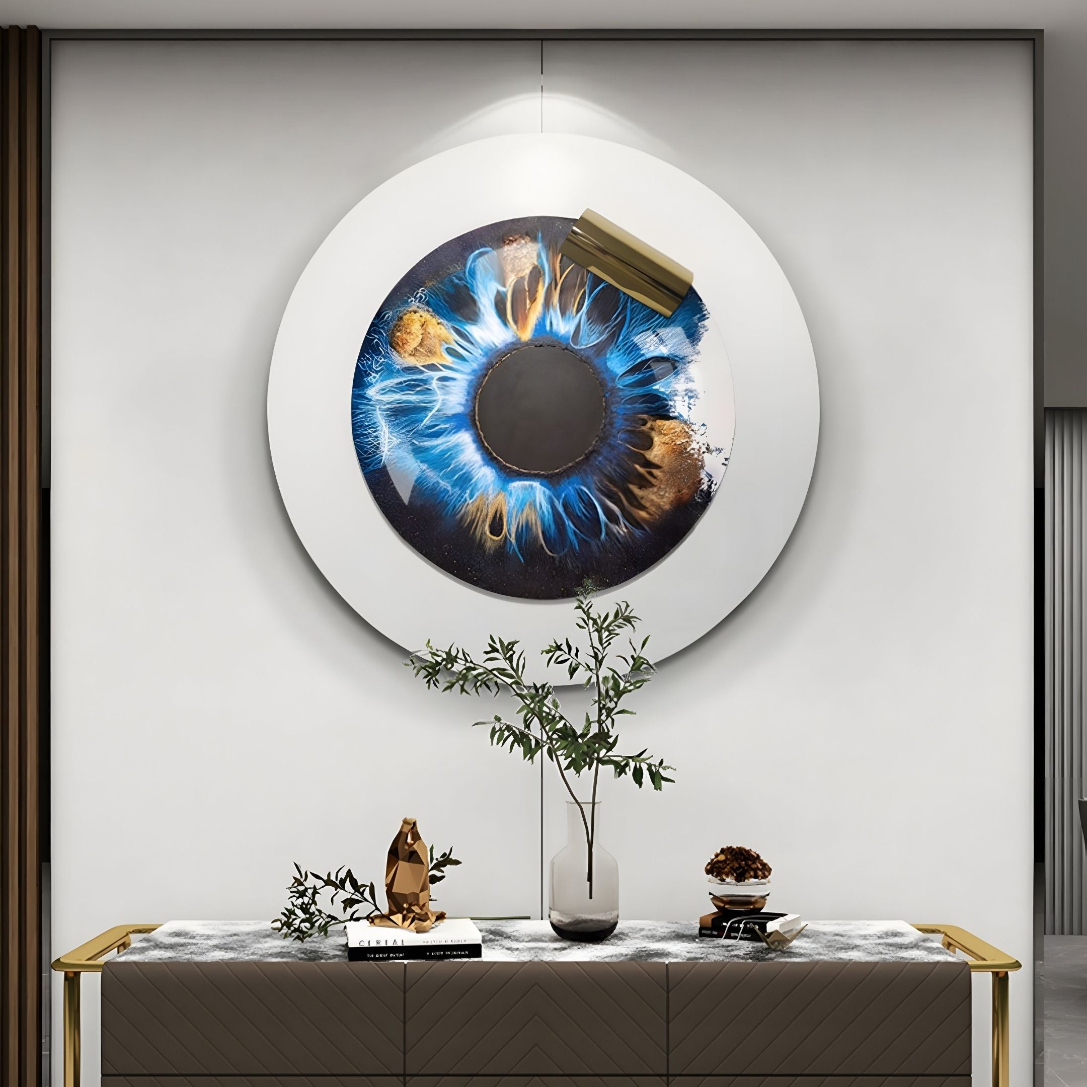 A modern interior displays the Iriselle Cosmic Blue Crystal Porcelain Round 3D Wall Art by Giant Sculptures, resembling an eye in blue and gold above a sideboard adorned with a vase of leafy branches, books, and decorative accents for designer flair.
