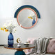 The Arcaluna Navy & Green Layered Round Mirror 3D Wall Art by Giant Sculptures hangs on a white wall above a modern living room with a glass table, blue vase of white and yellow flowers, pink tufted bench, and gray throw blanket.
