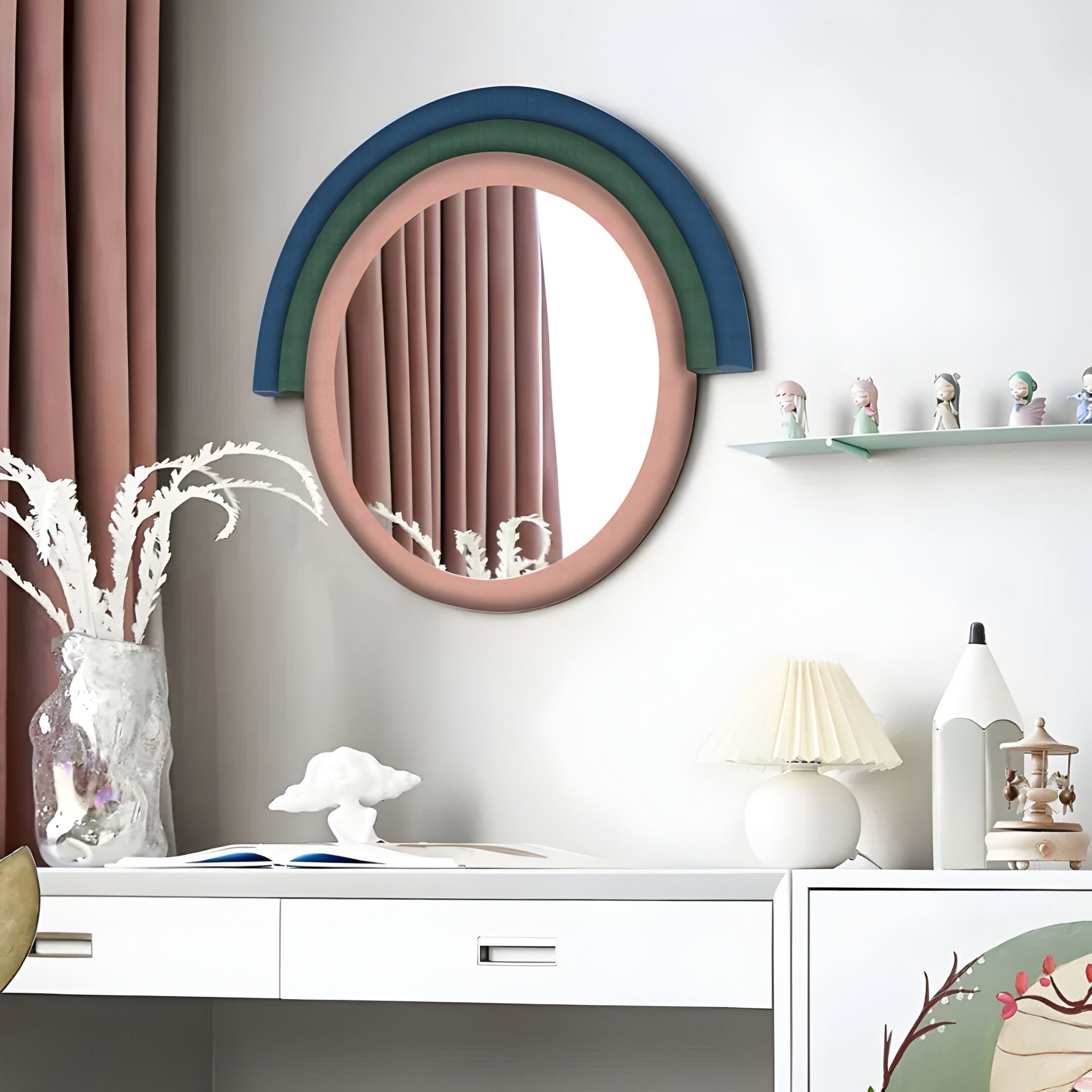 A modern desk with a white surface, lamp, decor, and a vase sits below the Arcaluna Navy & Green Layered Round Mirror 3D Wall Art by Giant Sculptures; pink curtains and nearby shelves complete the look.