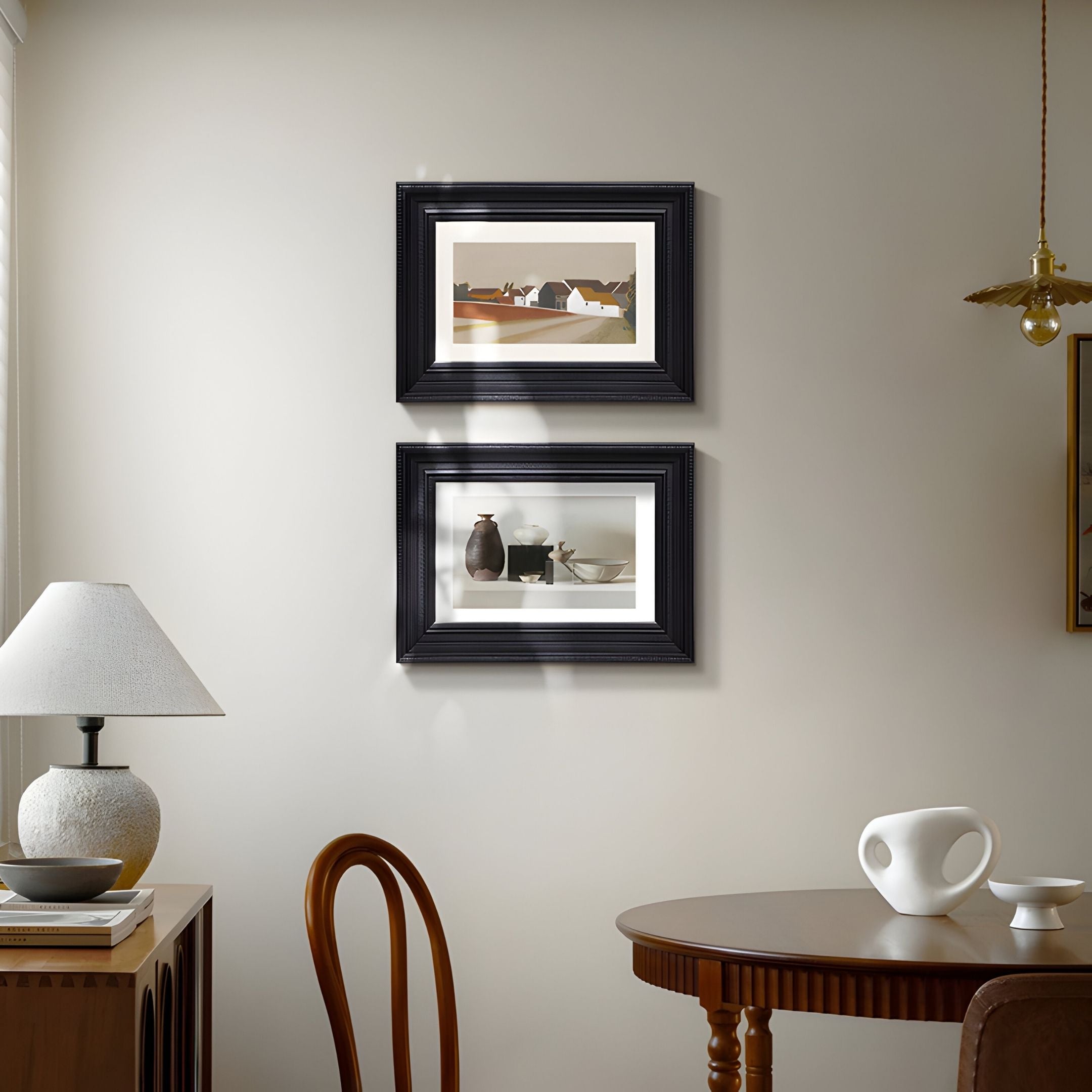 A cozy dining room with a round table, chairs, and side table glows with natural light. Above the beige wall, the Ardensir Village & Still Life Pottery 2-Set Framed Wall Art from Giant Sculptures adds vintage charm and style.