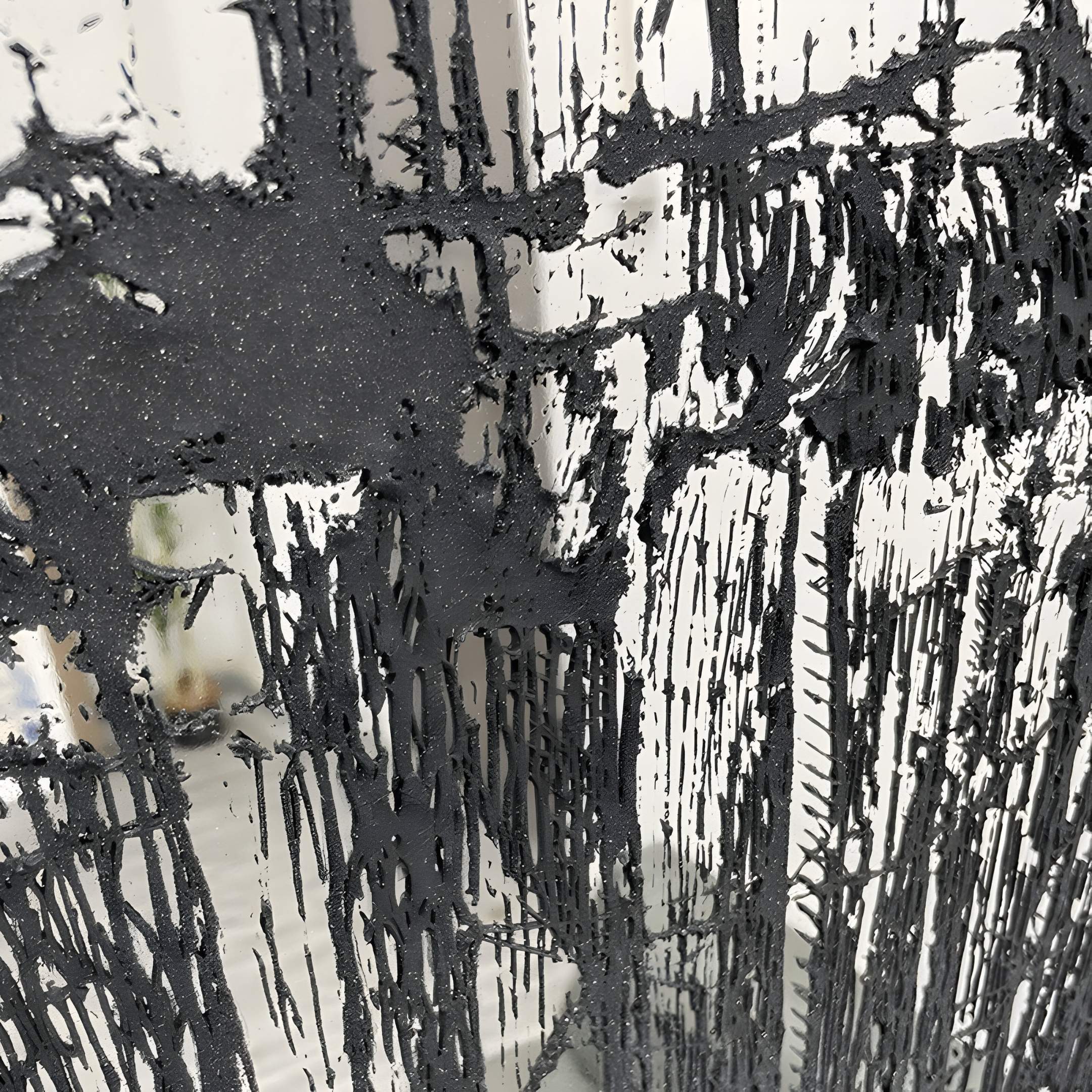The Rivarenne Black Abstract Texture Steel Mirror 3D Wall Art by Giant Sculptures features vertical streaks inspired by dripping paint or ink on glass, creating a bold, uneven pattern that stands out against a blurred light background.