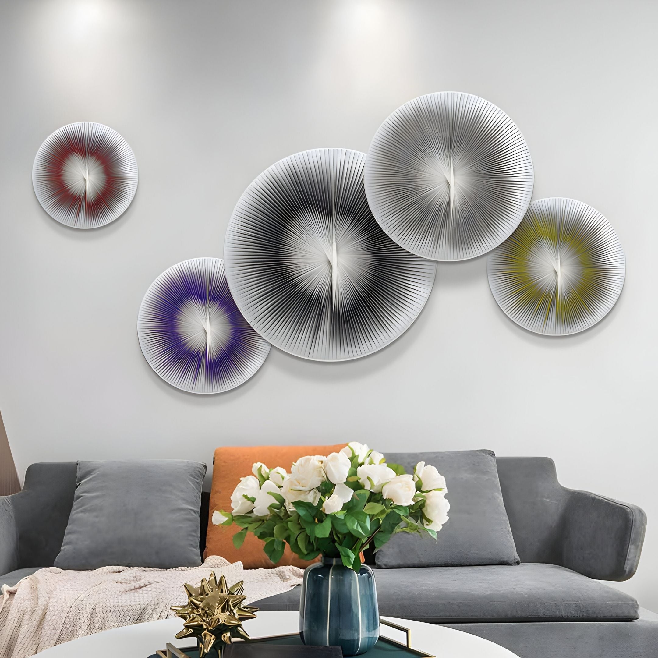 A modern living room with a gray sofa, orange cushion, and white flower arrangement on a round table features the Centria II Multicolour Disc 5-Panel 3D Leather Wall Art by Giant Sculptures—five abstract discs in black, red, purple, yellow, and white.