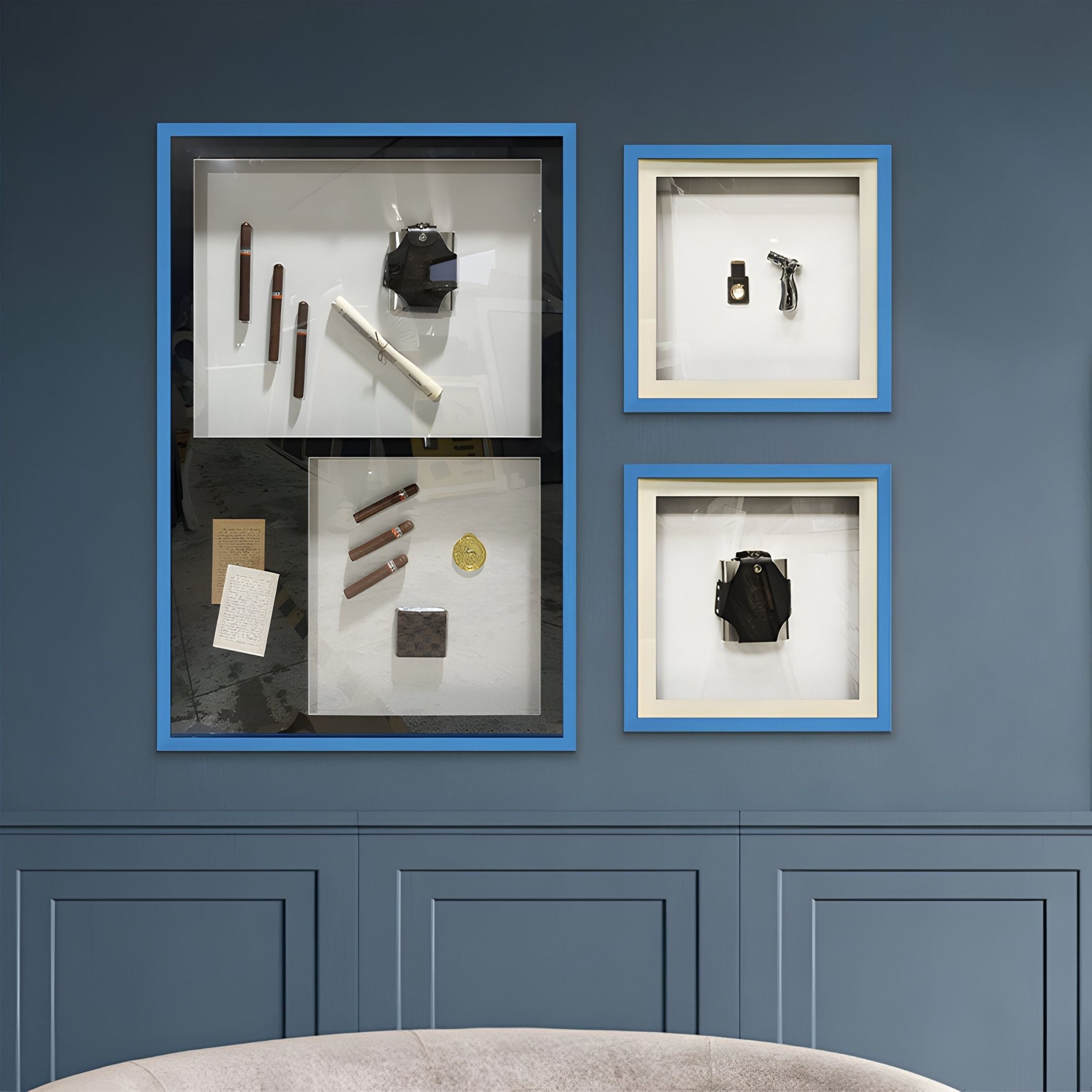 Three framed display cases of contemporary still life, including the Valet Noir Cigar Cutter & Torch Blue Framed Wall Art by Giant Sculptures, are mounted on a dark blue wall above a light gray sofa.