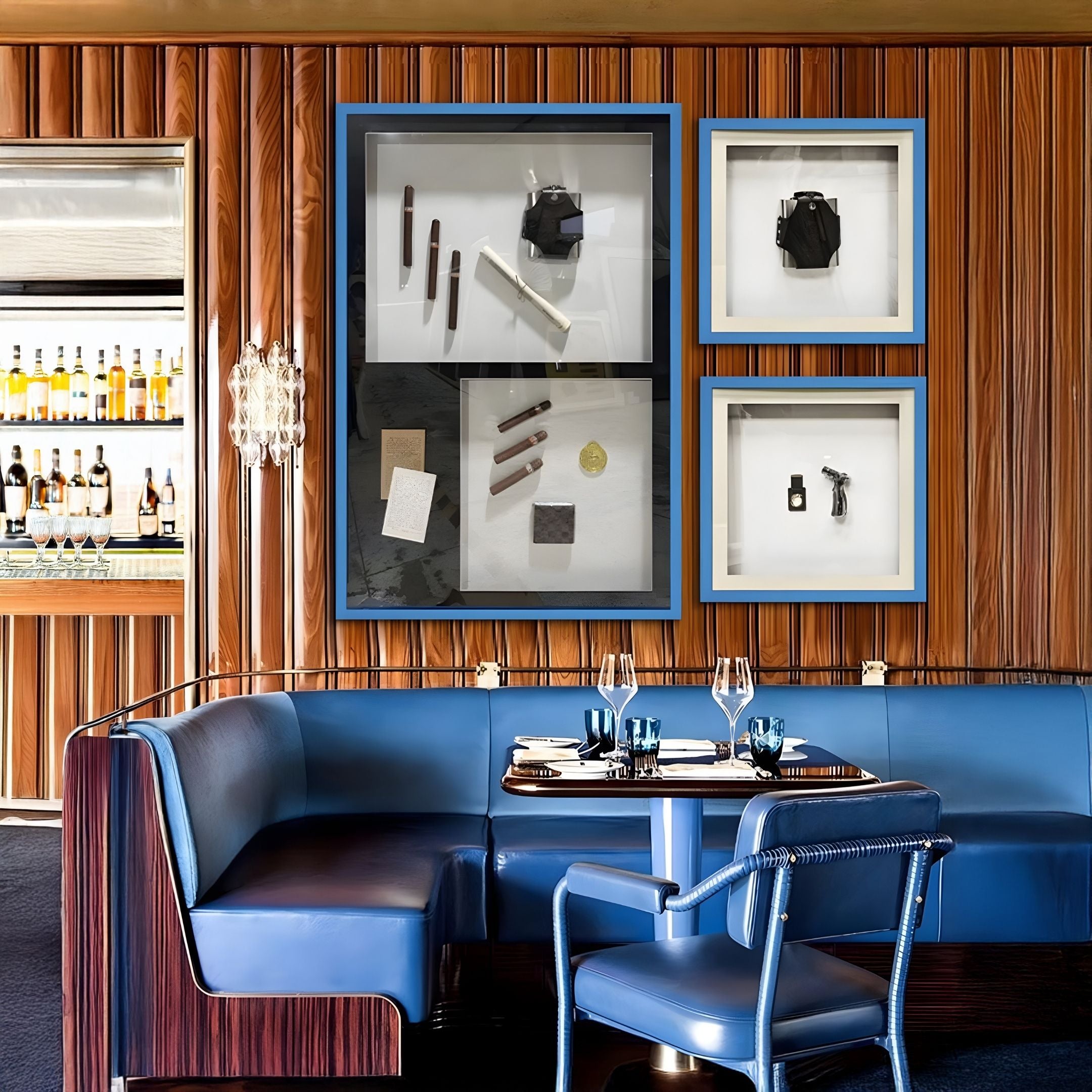 A stylish restaurant interior features wood-paneled walls, a blue leather booth and chairs, set tables, and Giant Sculptures' Valet Noir Leather Flask Blue Framed Wall Art. A bar with bottles appears in the background.