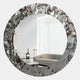 The Aurargent Round Layered Silver Mirror 3D Wall Art by Giant Sculptures features a textured metallic frame, reflecting a modern living room with a grey sofa, white coffee table, minimalist decor, and large white curtains in natural light.