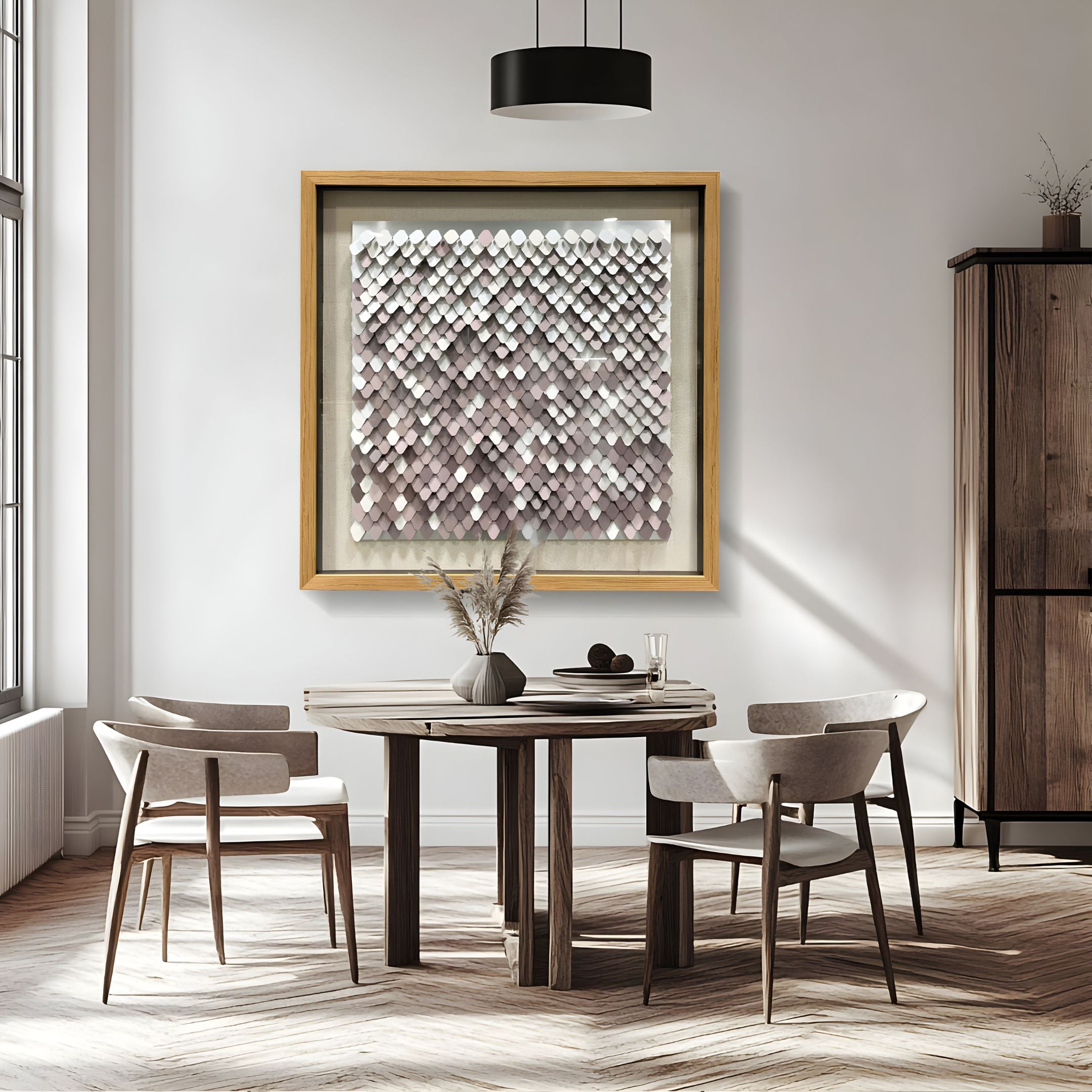 A modern dining room with a round wooden table, four light upholstered chairs, a vase of dried plants, and the Tessora White & Blush Scale Mosaic Metal 3D Wall Art by Giant Sculptures in a gold frame on neutral walls and wood flooring.