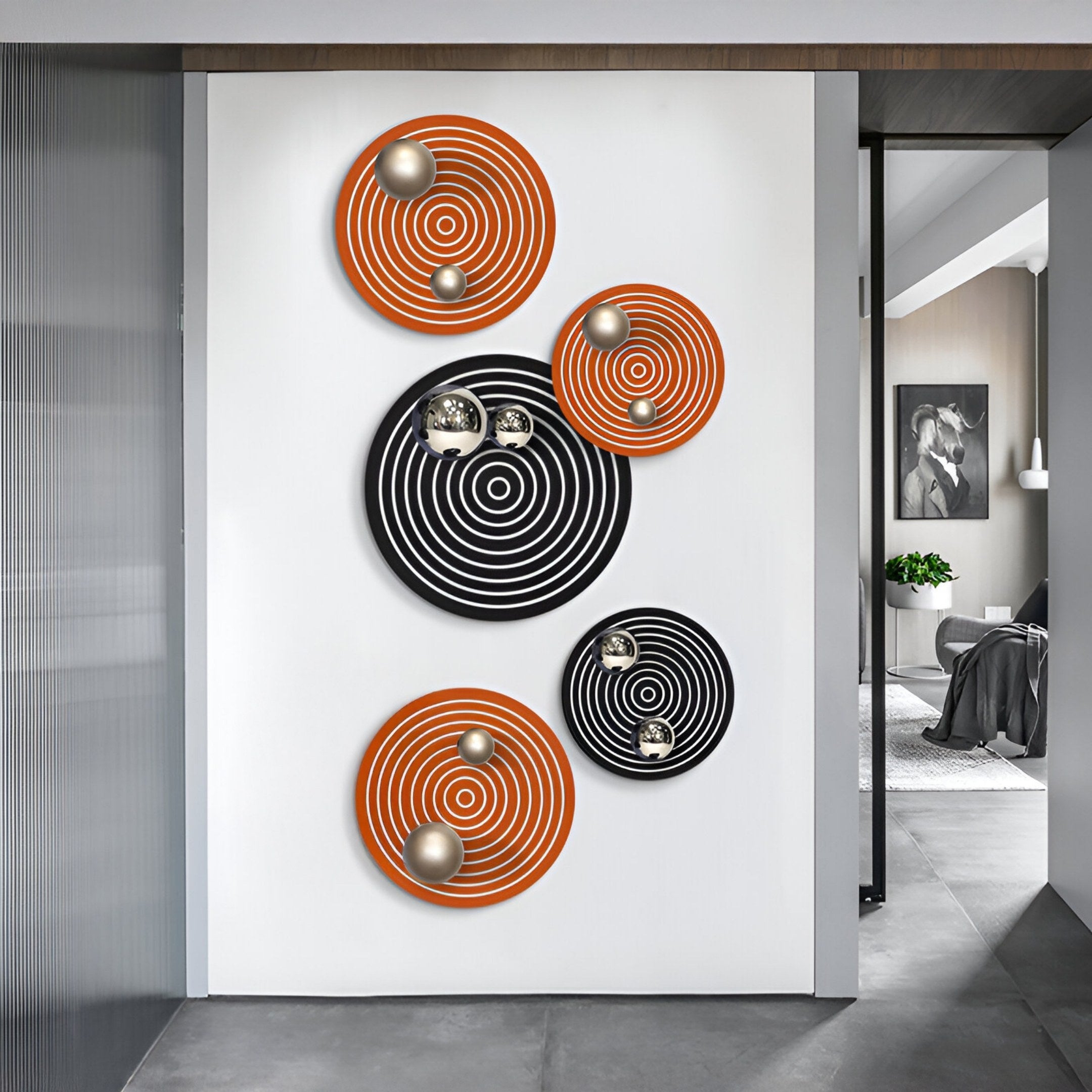 Orbiora Orange & Black 5-Panel Concentric Sphere 3D Wall Art by Giant Sculptures features three orange and two black concentric circles with metallic spheres, creating striking 3D decor mounted on a white wall in a modern interior. A living area is visible in back.