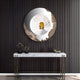 The Portara White Cloister Illusion Round 3D Wall Art by Giant Sculptures, featuring an abstract metallic edge, hangs above a white marble console on a dark striped wall. The mirror reflects an arched hallway; decor and a small box rest below.