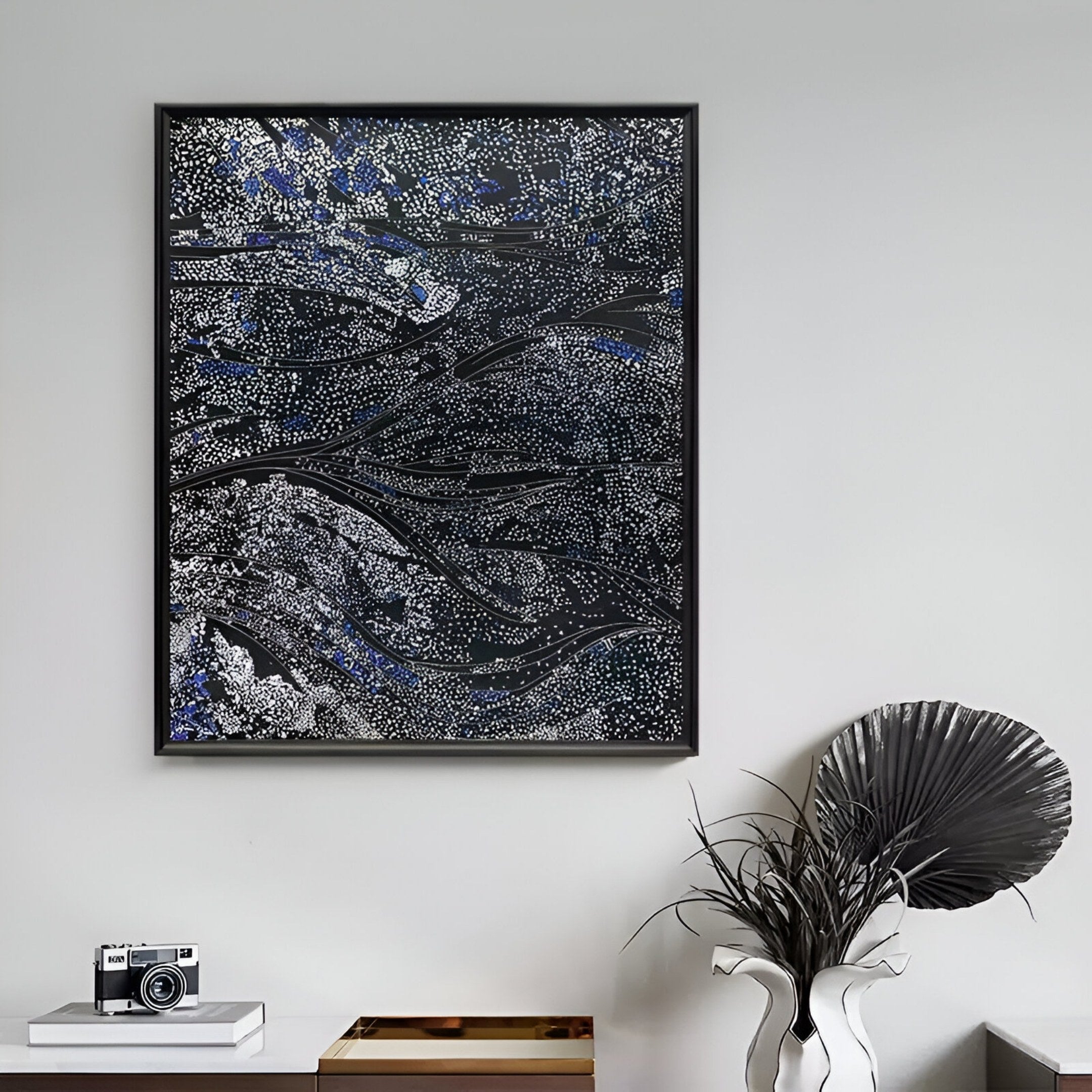 The Noctyra Midnight Flow Abstract 3D Wall Art by Giant Sculptures, featuring swirling black, white, and blue patterns, hangs on a light gray wall above a white table with a potted plant, tray, and vintage camera.