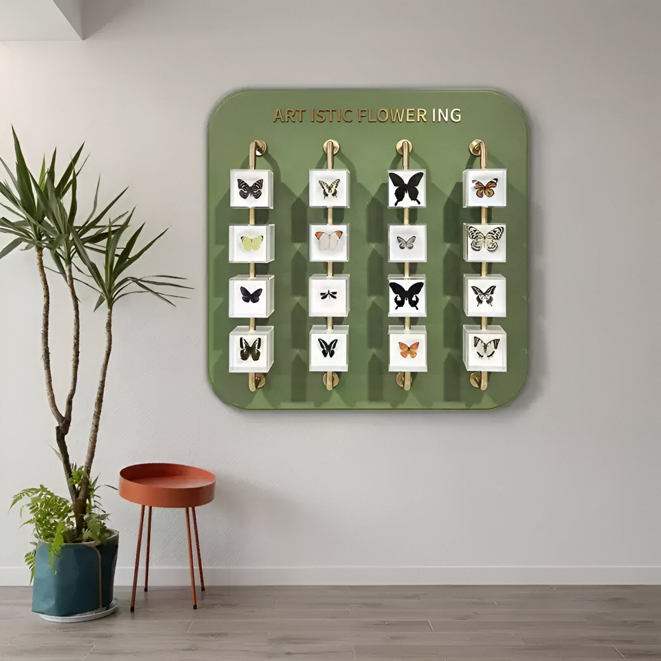 The Aurelian Butterfly Display Acrylic 3D Wall Art by Giant Sculptures features twelve framed butterflies on a green wall, gold letters spelling ARTISTIC FLOWERING (some missing), plus a potted plant and red chair for museum-inspired style.