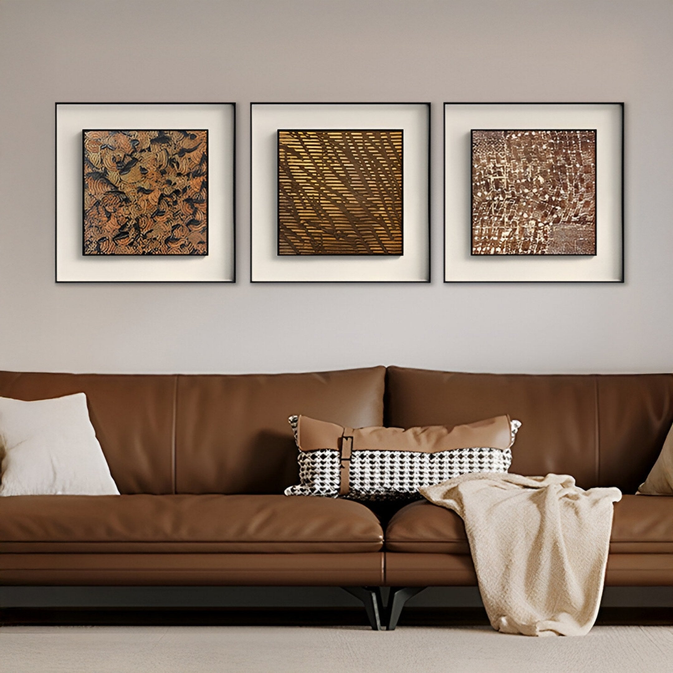 A brown leather sofa with a cream throw and patterned cushion sits against a beige wall. Above, three Fiorenze Copper & Black Swirl Textured Square 3D Wall Art pieces by Giant Sculptures display earthy tones and textured patterns.