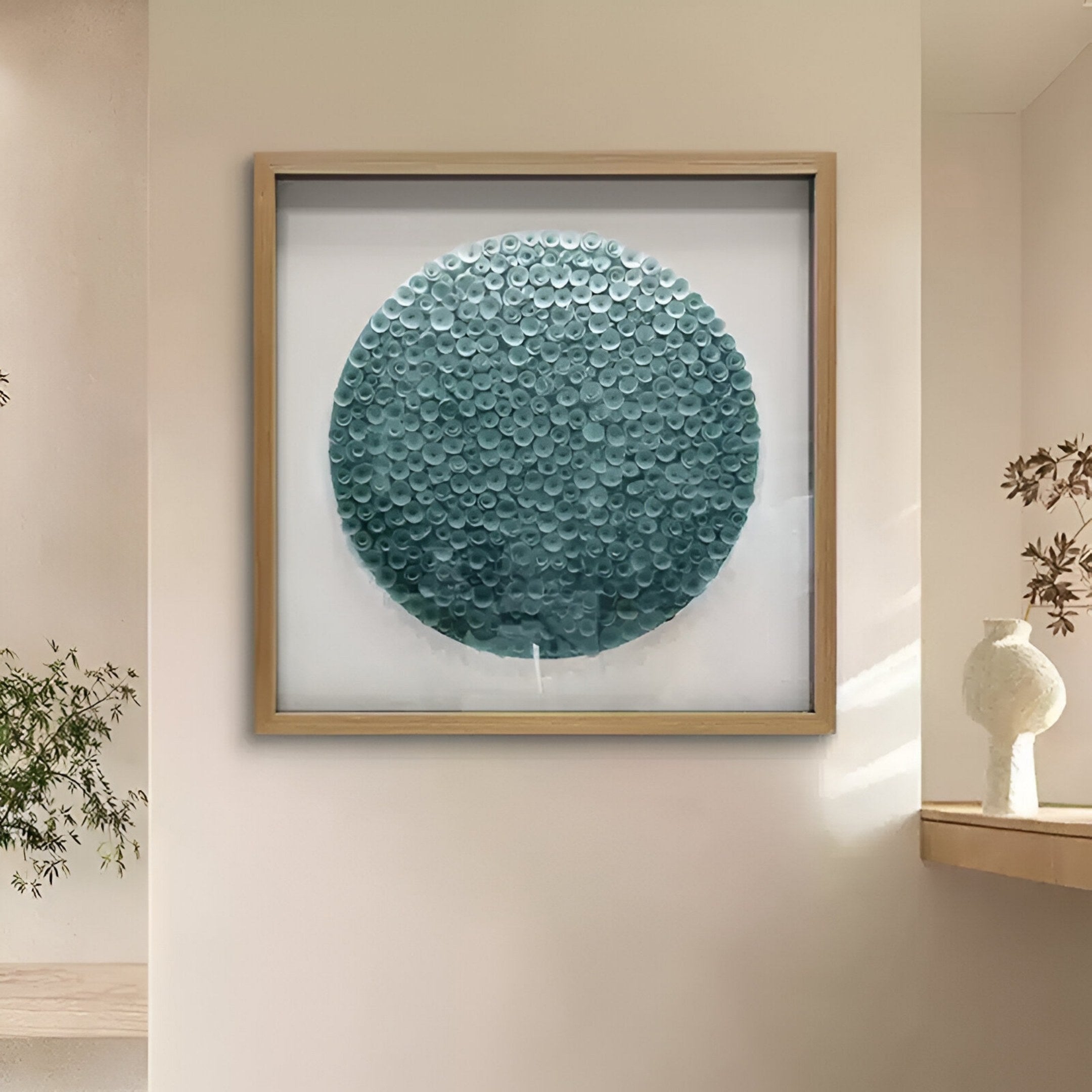 The Aureliare Teal Blue Rolled Floral Framed 3D Wall Art by Giant Sculptures, featuring a textured circular pattern, is displayed on a light wall near a leafy plant and a beige vase with dried stems on a wooden shelf.