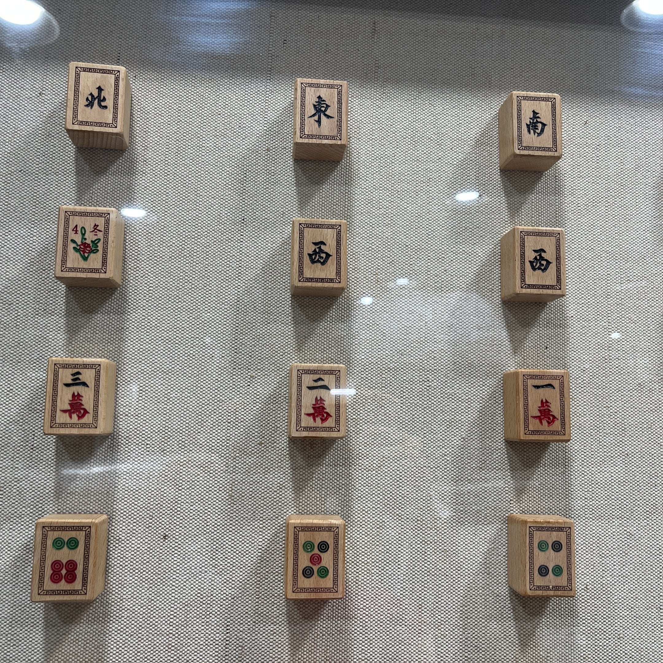 Close detail of the Fortuna Mahjong Carved Wood Tile Linen Wall Art, showing carved honour tiles for the winds alongside character and circle suit tiles on the woven linen ground.