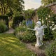 Winged Angel Boy Marble Outdoor Sculpture – 80cm by Giant Sculptures, a white marble cherub in an animated pose holding a bow, set in a lush walled garden with delphiniums and roses.