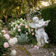 Little Angels Marble Outdoor Sculpture by Giant Sculptures, 80cm white marble winged angel boy playing a violin, set in a lush garden with pink peonies and climbing roses.