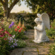 Praying Angel Marble Outdoor Sculpture by Giant Sculptures, a 70cm white marble kneeling angel with clasped hands and detailed wings, set in a garden with colorful flowers and a stone wall.