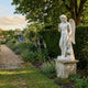Famous Cupid Art Angel Marble Outdoor Sculpture in white natural marble, 160cm, shown in a lush English walled garden beside flowering borders and a clipped yew hedge.