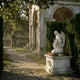 Eros And Snake Cupid Angel Marble Outdoor Sculpture by Giant Sculptures, 100cm white marble figure seated on a pedestal with wings and serpent, set in a garden with stone arches and hedges.