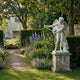 Eros God of Love Angel Marble Outdoor Sculpture by Giant Sculptures, 130cm white marble figure with wings drawing a bow, displayed on a stone pedestal in a lush garden.