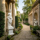 Giant Sculptures Cupid God Of Love Angel Marble Outdoor Sculpture in white marble, shown in a classical garden setting with stone arches, greenery, and a gravel path.