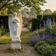 Standing Angel Marble Outdoor Sculpture by Giant Sculptures, 150cm white marble angel holding flowers on a square pedestal, set in a lush English garden with delphiniums, foxgloves, and lavender.