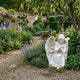 Kneeling Angel Marble Outdoor Sculpture by Giant Sculptures, 80cm white marble figure with crossed arms and detailed wings on a gravel path in a walled garden with roses and lavender.