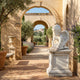 Small Angel Reading Angel Marble Outdoor Sculpture by Giant Sculptures, a 50cm white marble cherub seated and reading a book on a square marble pedestal, displayed in a sunlit arcaded courtyard with olive trees and lavender.
