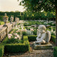 Sleeping Baby Angel Marble Outdoor Sculpture by Giant Sculptures, seated with head resting on one hand, wings folded, in a formal garden with boxwood hedges and pink peonies.