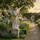Large Angel Marble Outdoor Sculpture by Giant Sculptures, 180cm white marble winged figure on a weathered stone pedestal beside a gravel path in a flowering walled garden.