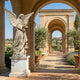 Large Angel Marble Outdoor Sculpture by Giant Sculptures, 180cm white marble winged angel on a pedestal beneath a stone arch with olive trees and lavender.
