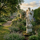 Four Season Angel white marble outdoor sculpture by Giant Sculptures shown in a formal garden setting amid flowering borders, standing on a square pedestal with wings, classical drapery, and floral attributes.