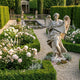 Ponte Sant Angelo V1 Angel Marble Outdoor Sculpture by Giant Sculptures, 180cm white marble figure in flowing robes with outstretched wings, displayed on a gravel path in a formal garden with roses and hedges.