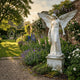 Classic Praying Angel Marble Outdoor Sculpture by Giant Sculptures, 180cm white marble angel with spread wings and flowing robes holding flowers, set in a lush English walled garden.