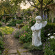Giant Sculptures Cherub V2 Angel Marble Outdoor Sculpture in white natural marble, 80cm tall, shown in an English cottage garden beside a gravel path with flowering borders and a brick wall.