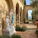 Lourdes Virgin Mary Angel Marble Outdoor Sculpture by Giant Sculptures, showing white marble Our Lady of Lourdes with teal sash beside kneeling Saint Bernadette on a shared base in a sunlit Mediterranean courtyard.