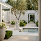 Winged Angel Marble Outdoor Sculpture by Giant Sculptures, 160cm white marble figure kneeling beside a reflective pool in a landscaped courtyard with olive trees and hydrangeas.