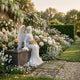 Giant Sculptures Cemetery Angel Marble Outdoor Sculpture – 150cm, a white marble winged angel seated on a dark stone pedestal holding orange roses, set in a lush garden border with pink peonies and climbing roses.