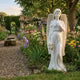 Angel Holding Flowers Marble Outdoor Sculpture – 160cm by Giant Sculptures, a white marble winged angel in flowing drapery holding a carved bouquet, displayed in a lush English garden with mixed flower borders and a gravel path.