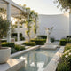 Garden Angel Marble Outdoor Sculpture by Giant Sculptures, 180cm white marble winged angel holding flowers on a stepped pedestal beside a reflecting pool in a landscaped courtyard.