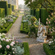 Kneeling Angel With Candlestick Marble Sculpture by Giant Sculptures, 80cm white marble angel with wings and flowing drapery, displayed in a formal garden with boxwood hedges and roses.
