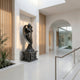 Black Angel Marble Outdoor Sculpture by Giant Sculptures, 160cm tall, depicting a winged angel in flowing robes with arms crossed on an ornate pedestal, displayed in a modern white hallway with arched doorway and greenery.