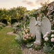 Angel With Large Wings marble outdoor sculpture by Giant Sculptures shown in an English cottage garden surrounded by pink roses and flowering borders at golden hour.