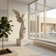 Life-Size Angel Marble Outdoor Sculpture by Giant Sculptures, 200cm white marble figure with outstretched wings and draped robes on a stone pedestal, shown in a modern interior beside floor-to-ceiling windows overlooking a pool courtyard.
