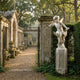 Winged Sitting Cherub Angel Marble Outdoor Sculpture in white marble on a pedestal beside a stone gateway in a classical garden setting with colonnade and mature trees.